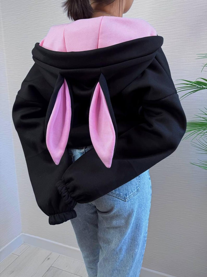 Op de afbeelding: Zwarte hoodie met roze konijnenoren en een roze binnenkant. De sweatshirt heeft lange mouwen met geribde manchetten. Het model draagt een blauwe spijkerbroek.