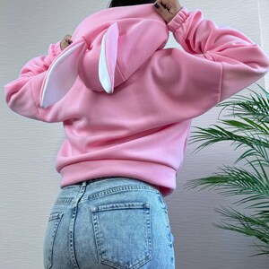 Op de afbeelding: Een lichtroze hoodie met een konijnenoordesign op de capuchon. De hoodie heeft lange mouwen en wordt gedragen met een blauwe jeans. De konijnenoren zijn wit met roze binnendetails.