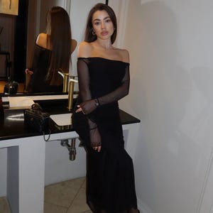 May include: A black off-the-shoulder midi dress with long sheer mesh sleeves, paired with black sheer tights and black pointed-toe high-heeled pumps. The dress features a straight neckline and a fitted silhouette that extends to the mid-calf.