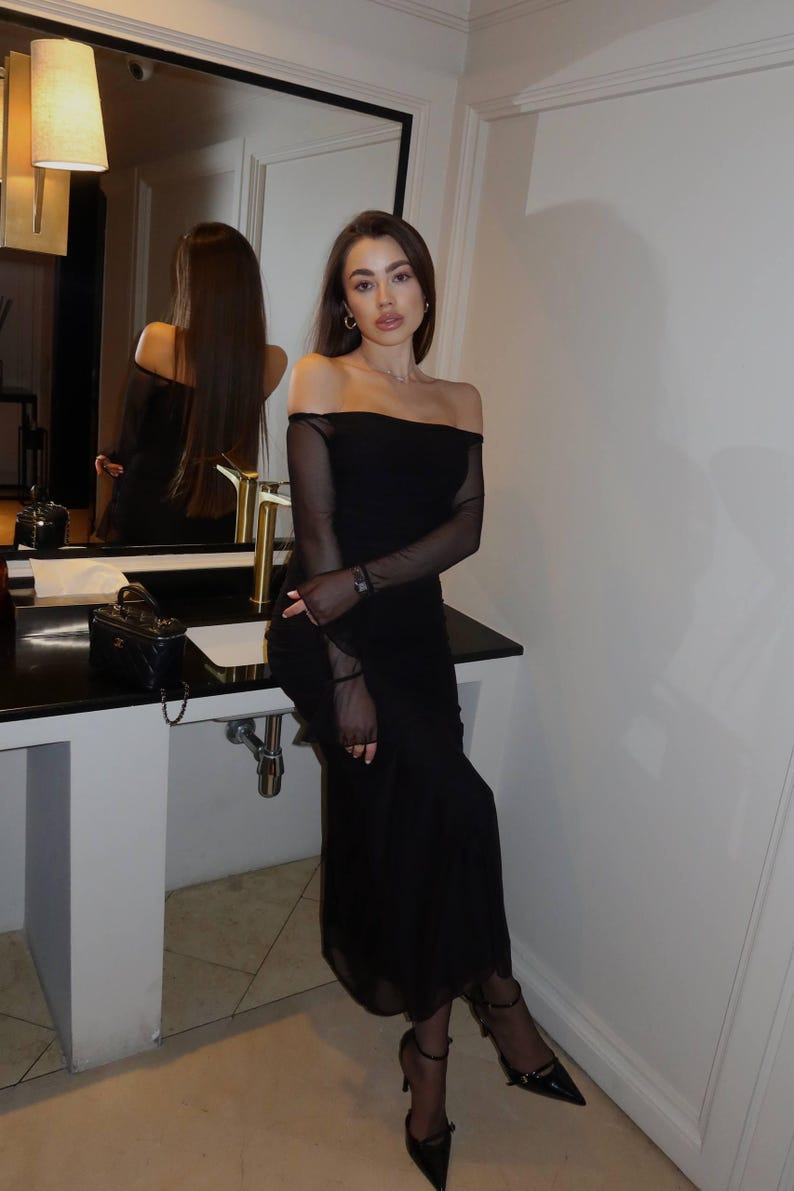 May include: A black off-the-shoulder midi dress with long sheer mesh sleeves, paired with black sheer tights and black pointed-toe high-heeled pumps. The dress features a straight neckline and a fitted silhouette that extends to the mid-calf.