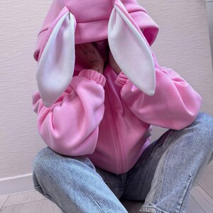 Op de afbeelding: Een roze hoodie met grote konijnenoren en een witte binnenkant. De hoodie heeft een ritssluiting en wordt gedragen met lichtblauwe jeans. De mouwen zijn bij de polsen gerimpeld.