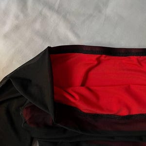 May include: A garment featuring a sheer black mesh outer layer over a solid red fabric lining. The piece has a wide, dark black elasticized waistband at the top edge, with visible stitching along the hem where the mesh meets the red base layer.