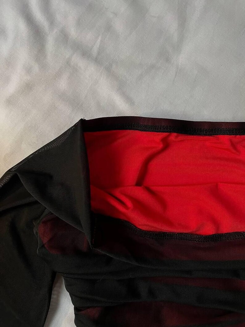 May include: A garment featuring a sheer black mesh outer layer over a solid red fabric lining. The piece has a wide, dark black elasticized waistband at the top edge, with visible stitching along the hem where the mesh meets the red base layer.