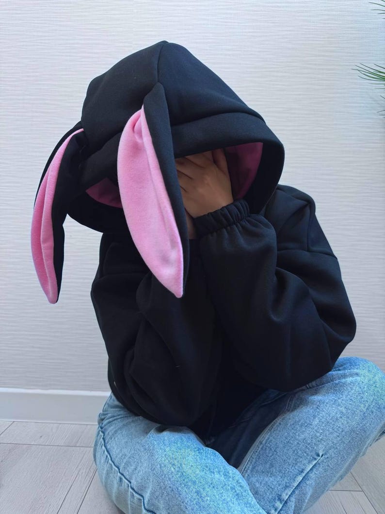 Op de afbeelding: Zwarte hoodie met een konijnenoordesign. De capuchon heeft lange, floppy oren met roze binnenvoering. De persoon op de foto draagt een blauwe spijkerbroek. De mouwen van de hoodie zijn bij de polsen gerimpeld.