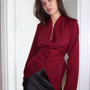 Op de afbeelding: Een bordeauxrode blazer met een V-hals en een sluiting met drie knopen. De jas met lange mouwen wordt gecombineerd met een zwarte minirok. De outfit wordt gedragen door een persoon in een deuropening.