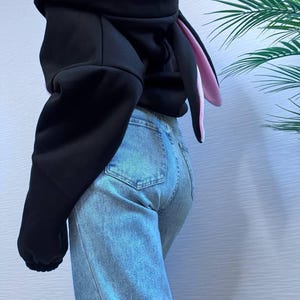 Op de afbeelding: Zwarte hoodie met roze voering en lange konijnenoren. De hoodie wordt gecombineerd met lichtblauwe jeans. De mouwen zijn lang en de capuchon is omhoog. De konijnenoren zijn aan de binnenkant roze en aan de buitenkant zwart.