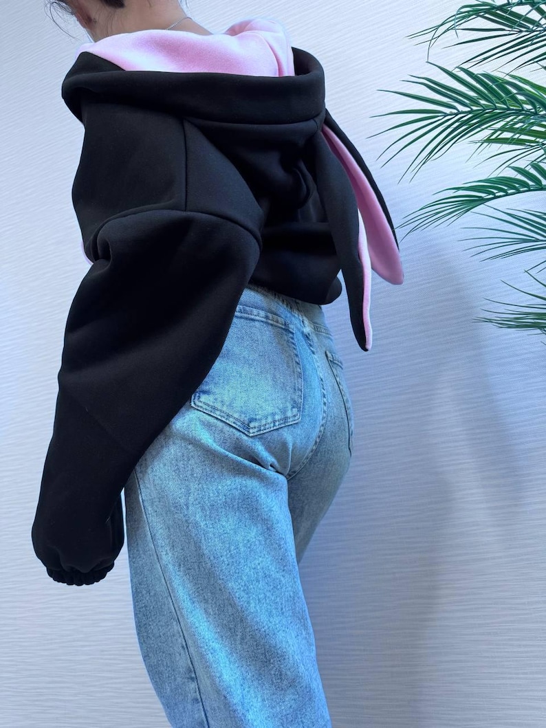 Op de afbeelding: Zwarte hoodie met roze voering en lange konijnenoren. De hoodie wordt gecombineerd met lichtblauwe jeans. De mouwen zijn lang en de capuchon is omhoog. De konijnenoren zijn aan de binnenkant roze en aan de buitenkant zwart.