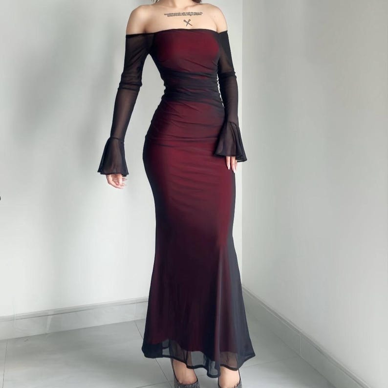 May include: A floor-length off-the-shoulder dress featuring a deep red base layer overlaid with sheer black mesh fabric. The garment includes long sleeves with flared cuffs, a fitted bodice, and a straight skirt silhouette.