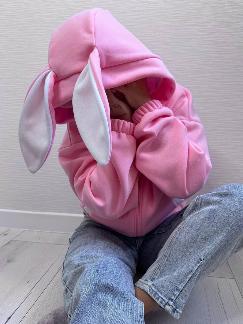 Op de afbeelding: Een roze konijnenhoodie met lange, slappe oren en een witte binnenkant. De hoodie heeft een ritssluiting en geribde manchetten. De persoon draagt een blauwe spijkerbroek. De outfit is casual en speels.