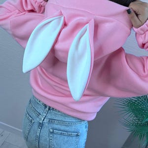 Op de afbeelding: Een roze hoodie met witte konijnenoren aan de capuchon. De sweatshirt heeft lange mouwen en een relaxte pasvorm. De persoon op de foto draagt een lichtblauwe spijkerbroek.