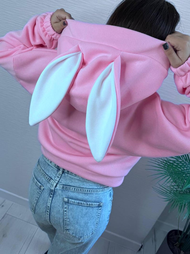 Op de afbeelding: Een roze hoodie met witte konijnenoren aan de capuchon. De sweatshirt heeft lange mouwen en een relaxte pasvorm. De persoon op de foto draagt een lichtblauwe spijkerbroek.