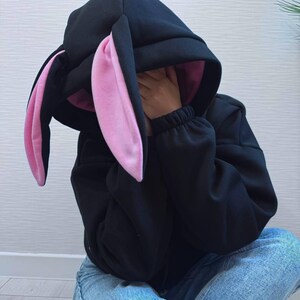 Op de afbeelding: Zwarte hoodie met een konijnenoordesign. De capuchon heeft lange, floppy oren met een roze binnenkant. De persoon op de foto draagt een blauwe spijkerbroek en zit. De mouwen van de hoodie zijn bij de polsen gerimpeld.