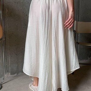 May include: A white midi-length skirt with a high-low hemline, featuring a gathered elastic waistband and a lightweight, semi-sheer crinkled fabric texture. The skirt has a loose, flowing silhouette that drapes down to the lower calf area.