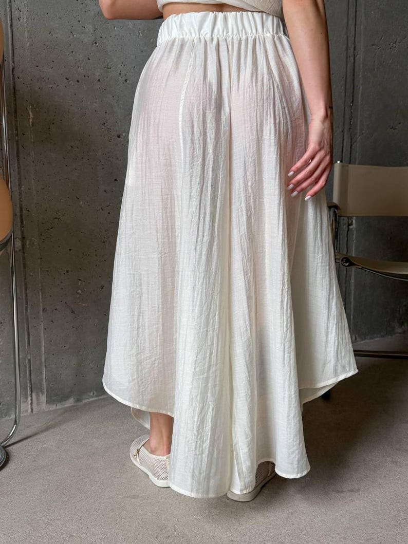 May include: A white midi-length skirt with a high-low hemline, featuring a gathered elastic waistband and a lightweight, semi-sheer crinkled fabric texture. The skirt has a loose, flowing silhouette that drapes down to the lower calf area.
