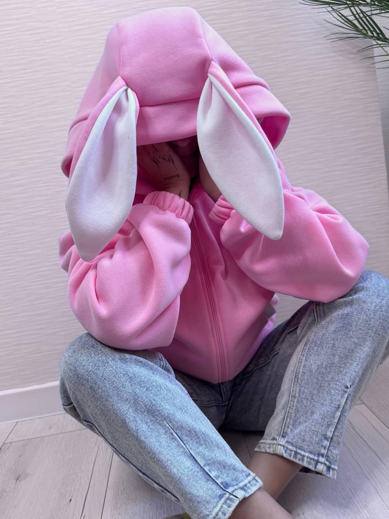 Op de afbeelding: Een roze hoodie met grote konijnenoren en een witte binnenkant. De hoodie heeft een ritssluiting en wordt gedragen met lichtblauwe jeans. De mouwen zijn bij de polsen gerimpeld.