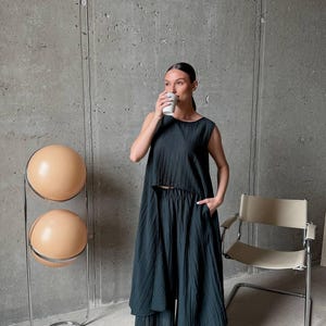 May include: A dark charcoal gray two-piece set consisting of a sleeveless, cropped, high-low top and wide-leg trousers. The fabric features a fine, vertical crinkled texture throughout, providing a lightweight and draped appearance for the garment.