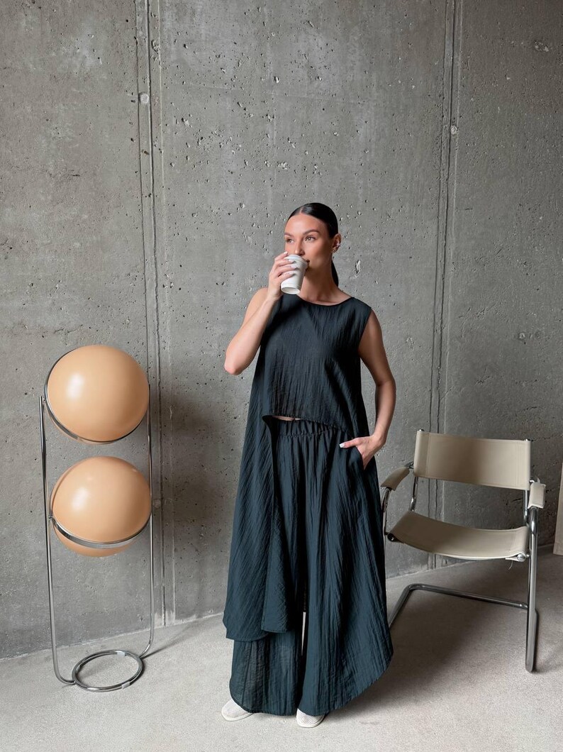 May include: A dark charcoal gray two-piece set consisting of a sleeveless, cropped, high-low top and wide-leg trousers. The fabric features a fine, vertical crinkled texture throughout, providing a lightweight and draped appearance for the garment.