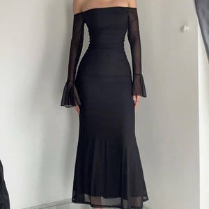 May include: A floor-length black dress featuring an off-the-shoulder neckline and long, sheer mesh sleeves that flare at the wrists. The garment has a fitted bodice that transitions into a flared, trumpet-style skirt with a sheer hemline.