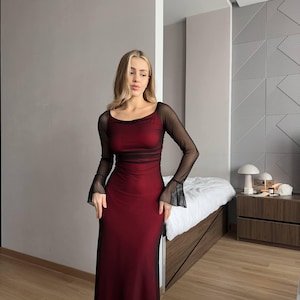 May include: A floor-length dress featuring a deep red base layer with a sheer black mesh overlay. The garment has a scoop neckline and long, fitted sleeves that flare slightly at the wrists, creating a layered, semi-sheer appearance.