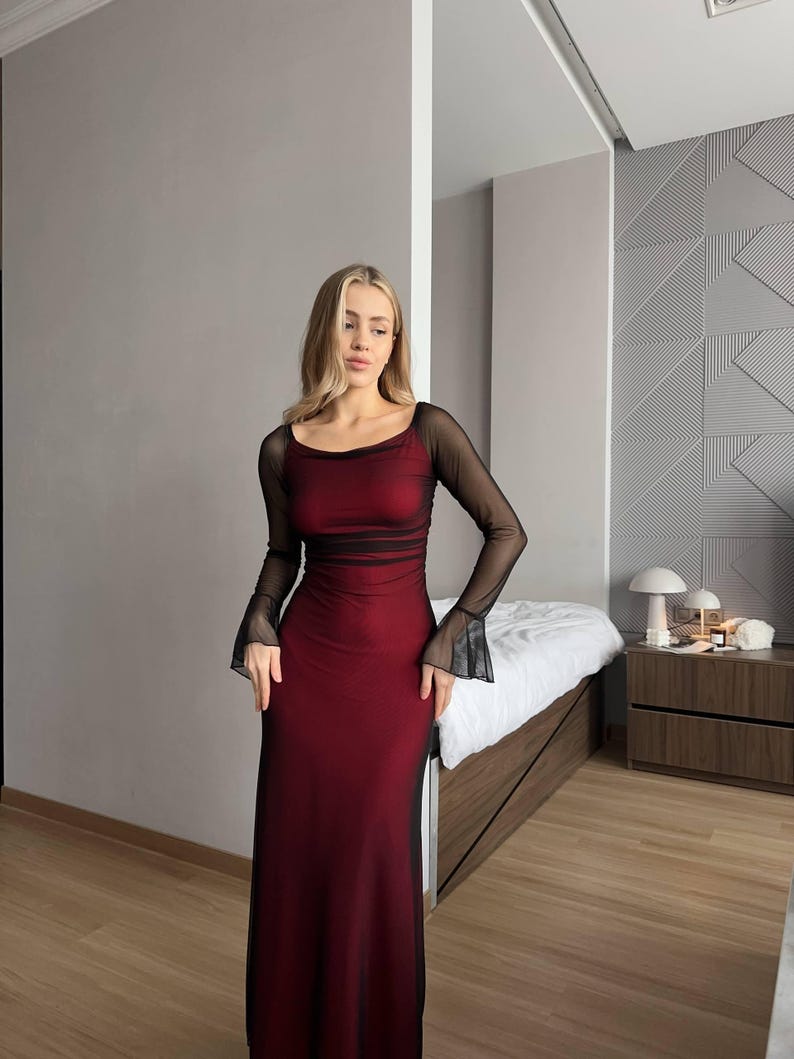 May include: A floor-length dress featuring a deep red base layer with a sheer black mesh overlay. The garment has a scoop neckline and long, fitted sleeves that flare slightly at the wrists, creating a layered, semi-sheer appearance.