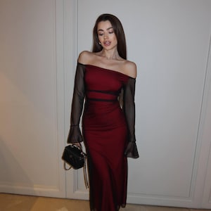 May include: A floor-length, off-the-shoulder dress in deep red with sheer black long sleeves and black mesh side panels. The garment features a straight neckline and a fitted silhouette. A small black structured handbag with a gold chain strap is held.