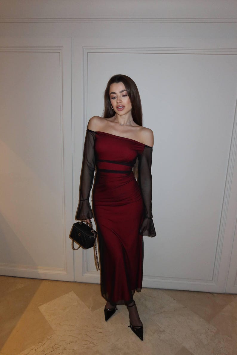 May include: A floor-length, off-the-shoulder dress in deep red with sheer black long sleeves and black mesh side panels. The garment features a straight neckline and a fitted silhouette. A small black structured handbag with a gold chain strap is held.