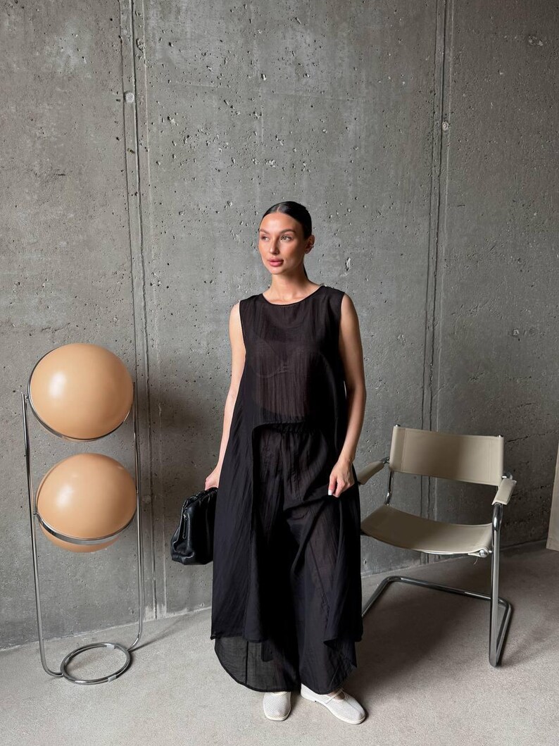 May include: A black sleeveless tunic top with a high-low hemline paired with matching wide-leg trousers. The outfit is made of a lightweight, semi-sheer fabric with a subtle vertical pleated texture. The top features a round neckline.