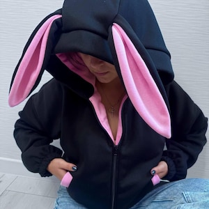 Op de afbeelding: Zwarte hoodie met konijnenoren. De capuchon heeft grote, roze gevoerde oren. De binnenvoering en de zakken zijn ook roze. De hoodie heeft een lange ritssluiting en wordt gedragen met een spijkerbroek.