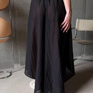 May include: A pair of black wide-leg trousers featuring a semi-sheer, textured fabric with vertical crinkled lines. The garment has an elasticized waistband and a loose, flowing silhouette that extends to a midi or maxi length hemline.