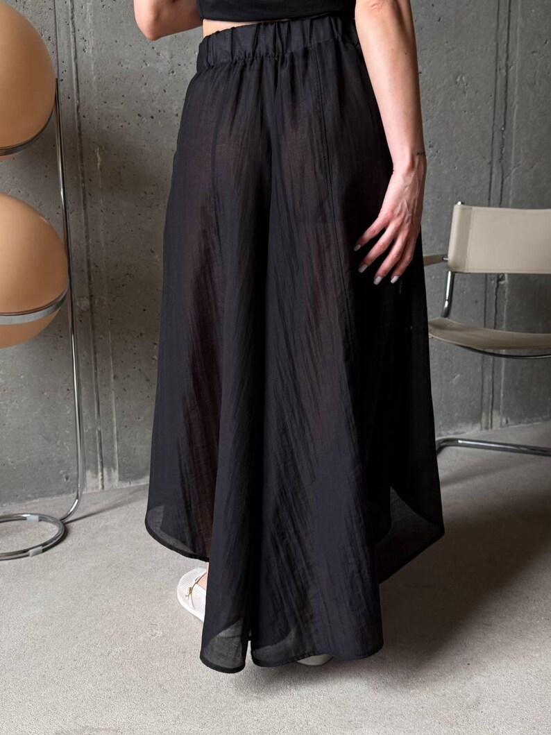 May include: A pair of black wide-leg trousers featuring a semi-sheer, textured fabric with vertical crinkled lines. The garment has an elasticized waistband and a loose, flowing silhouette that extends to a midi or maxi length hemline.