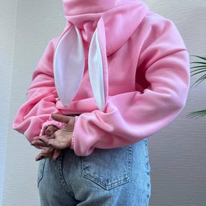 Op de afbeelding: Een roze hoodie met bevestigde konijnenoren. De oren zijn wit aan de binnenkant en roze aan de buitenkant. De sweatshirt is effen roze van kleur en heeft lange mouwen. De persoon draagt een blauwe spijkerbroek.