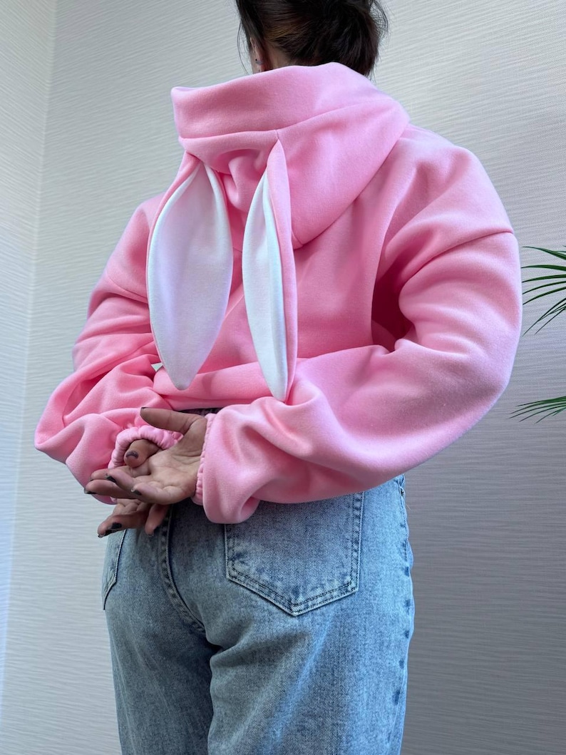 Op de afbeelding: Een roze hoodie met bevestigde konijnenoren. De oren zijn wit aan de binnenkant en roze aan de buitenkant. De sweatshirt is effen roze van kleur en heeft lange mouwen. De persoon draagt een blauwe spijkerbroek.