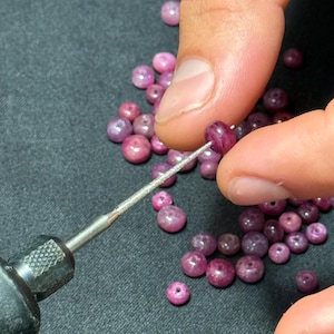 Może przedstawiać: Zbliżenie na kolekcję małych, okrągłych, różowo-czerwonych koralików z kamieni szlachetnych. Narzędzie jest włożone w jeden koralik, a inne koraliki są rozrzucone na ciemnej powierzchni. Koraliki wydają się być wywiercone do nawlekania, co sugeruje tworzenie biżuterii.