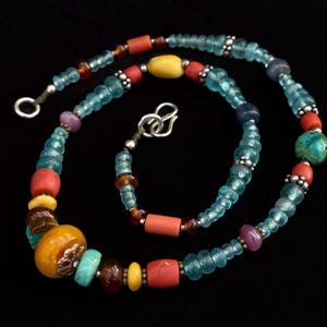Puede incluir: Un collar de cuentas colorido con una variedad de formas y tamaños. El collar presenta cuentas de color azul claro, rojo, amarillo y naranja, con detalles plateados. El collar se muestra sobre un fondo negro.