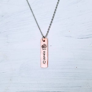 May include: A rectangular copper pendant necklace featuring a stamped tree design and the word SISU vertically aligned. The pendant hangs from a thin silver-toned metal chain, showcasing a metallic finish with clear black lettering and a detailed tree icon.