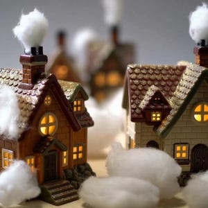 May include: A collection of miniature ceramic houses with illuminated windows and chimneys. The houses have brown, beige, and green roofs, with white cotton simulating snow. Fluffy cotton clouds are scattered around the houses, creating a winter village scene.