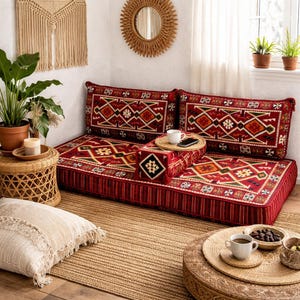 Peut inclure: Un canapé de sol bas, rouge, avec des coussins à motifs et un traversin assorti. Les coussins présentent un motif géométrique dans des tons de rouge, d'orange et de vert. Le canapé est posé sur un tapis tissé, créant un espace de vie bohème et confortable.