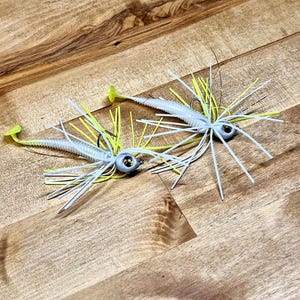 May include: Two fishing lures featuring light gray jig heads with dark eyes, white ribbed soft plastic bodies with paddle tails, and a skirt composed of thin, flexible white and neon yellow silicone strands extending outward from the head.