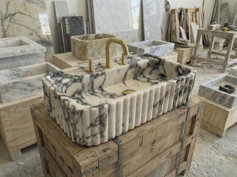Puede incluir: Un lavabo rectangular de m&aacute;rmol con un grifo dorado. El lavabo tiene un patr&oacute;n veteado blanco y gris y un borde acanalado. Se encuentra sobre una caja de madera, con otros lavabos en el fondo.
