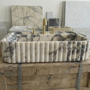 Puede incluir: Un lavabo rectangular de m&aacute;rmol con exterior estriado y grifos dorados. El lavabo presenta un patr&oacute;n veteado blanco y gris. Se exhibe sobre una caja de madera, con otras losas de m&aacute;rmol al fondo.
