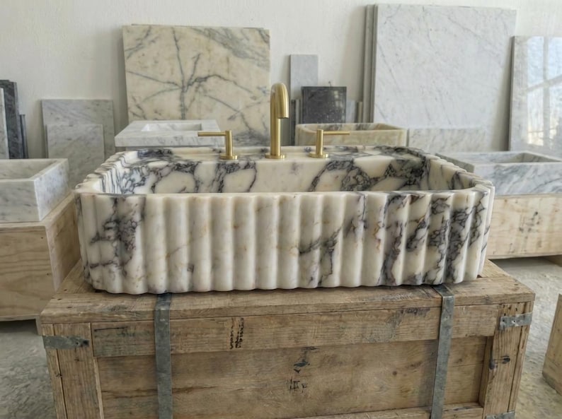 Puede incluir: Un lavabo rectangular de m&aacute;rmol con exterior estriado y grifos dorados. El lavabo presenta un patr&oacute;n veteado blanco y gris. Se exhibe sobre una caja de madera, con otras losas de m&aacute;rmol al fondo.
