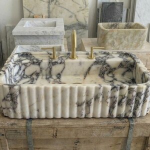 Puede incluir: Un lavabo rectangular de m&aacute;rmol con exterior acanalado y grifos dorados. El lavabo presenta un patr&oacute;n veteado blanco y gris. Otros lavabos de m&aacute;rmol son visibles en el fondo, mostrando una variedad de dise&ntilde;os.