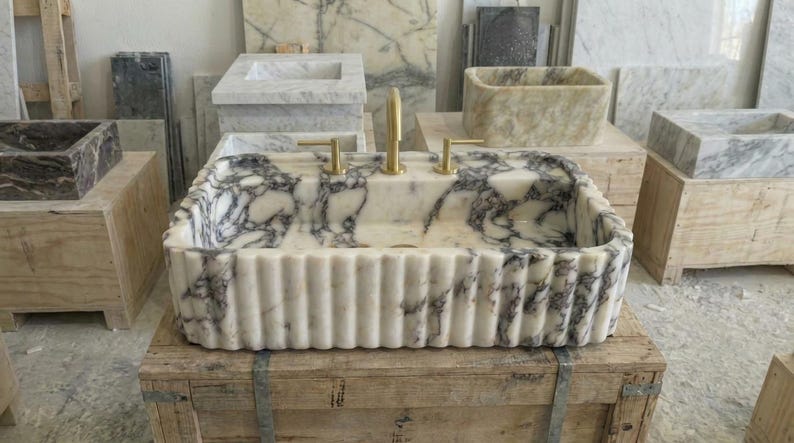 Puede incluir: Un lavabo rectangular de m&aacute;rmol con exterior acanalado y grifos dorados. El lavabo presenta un patr&oacute;n veteado blanco y gris. Otros lavabos de m&aacute;rmol son visibles en el fondo, mostrando una variedad de dise&ntilde;os.