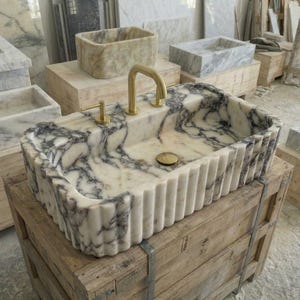 Puede incluir: Un lavabo rectangular de m&aacute;rmol con exterior estriado y grifo dorado. El lavabo presenta un patr&oacute;n veteado blanco y gris. Otros lavabos de m&aacute;rmol son visibles en el fondo, junto con cajas de madera.