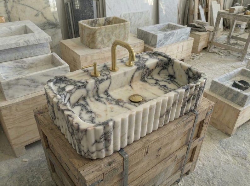 Puede incluir: Un lavabo rectangular de m&aacute;rmol con exterior estriado y grifo dorado. El lavabo presenta un patr&oacute;n veteado blanco y gris. Otros lavabos de m&aacute;rmol son visibles en el fondo, junto con cajas de madera.