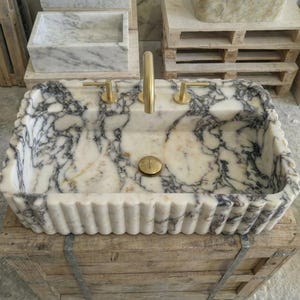 Puede incluir: Un lavabo rectangular de m&aacute;rmol con un patr&oacute;n veteado blanco y gris. El lavabo tiene un borde festoneado y grifos y accesorios dorados. El lavabo se exhibe sobre una caja de madera.