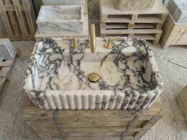 Puede incluir: Un lavabo rectangular de m&aacute;rmol con un patr&oacute;n veteado blanco y gris. El lavabo tiene un borde festoneado y grifos y accesorios dorados. El lavabo se exhibe sobre una caja de madera.