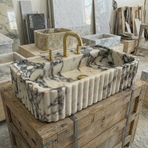 Puede incluir: Un lavabo rectangular de m&aacute;rmol con un grifo dorado. El lavabo tiene un patr&oacute;n veteado blanco y gris y un exterior estriado. Se encuentra sobre una caja de madera en un taller.
