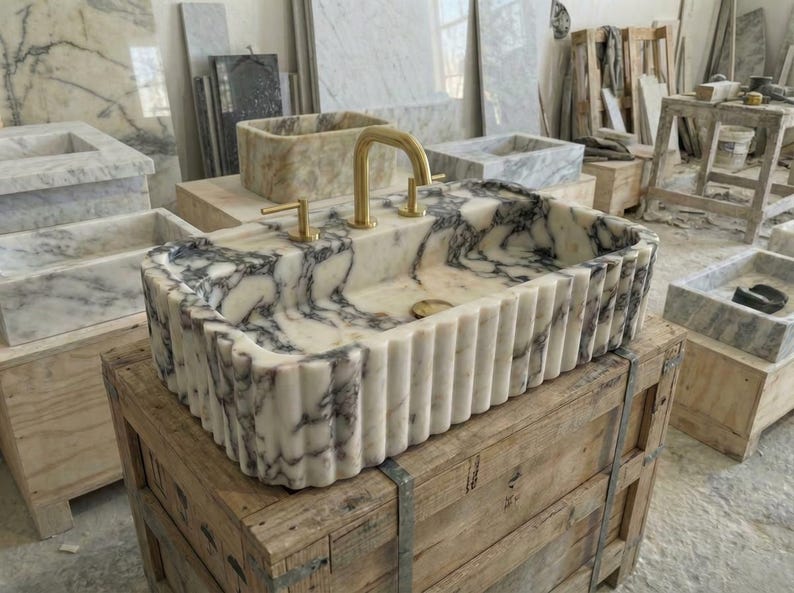 Puede incluir: Un lavabo rectangular de m&aacute;rmol con un grifo dorado. El lavabo tiene un patr&oacute;n veteado blanco y gris y un exterior estriado. Se encuentra sobre una caja de madera en un taller.