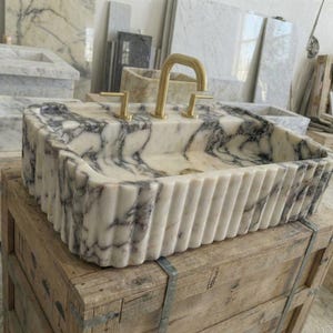 Puede incluir: Un lavabo rectangular de m&aacute;rmol con un patr&oacute;n veteado blanco y gris. El lavabo tiene un exterior acanalado y grifos dorados. El lavabo se exhibe sobre una caja de madera en un taller.